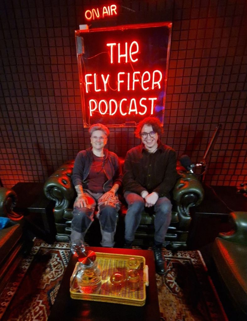 Vicky and Ross from the treehouse team sit in the Fly Fifer podcast recording studio, its a darkened room with comfortable leather seats, sound proofing on the walls and a neon sign behind saying " on air, the Fly Fifer podcast "